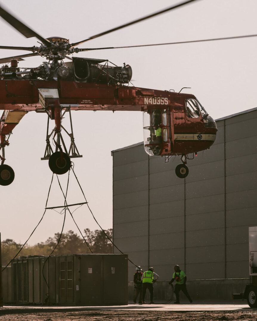 Construction workers attaching rigging to a massive HVAC unit under the Sikorsky S-64 helicopter.