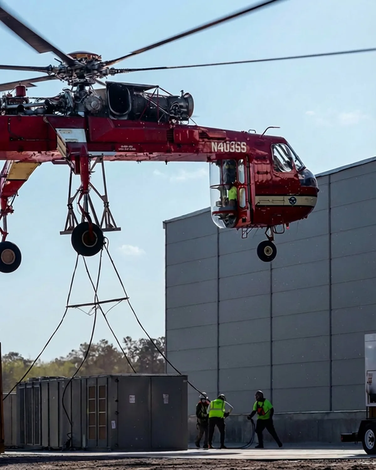 Construction workers attaching rigging to a massive HVAC unit under the Sikorsky S-64 helicopter.