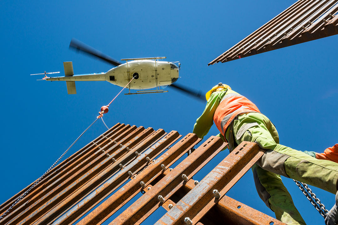 A white and blue Airbus AS350 (H125) helicopter hovering overhead, lowering a steel beam via a long-line to a construction worker standing on a high-rise steel structure under a clear blue sky.