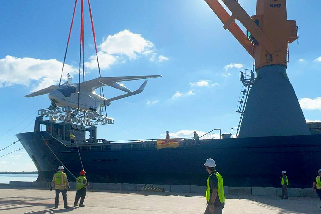 crane in mid-operation suspends Beta Technologies' ALIA CX300 electric aircraft above the docks at Port of Wilmington during transfer to cargo ship