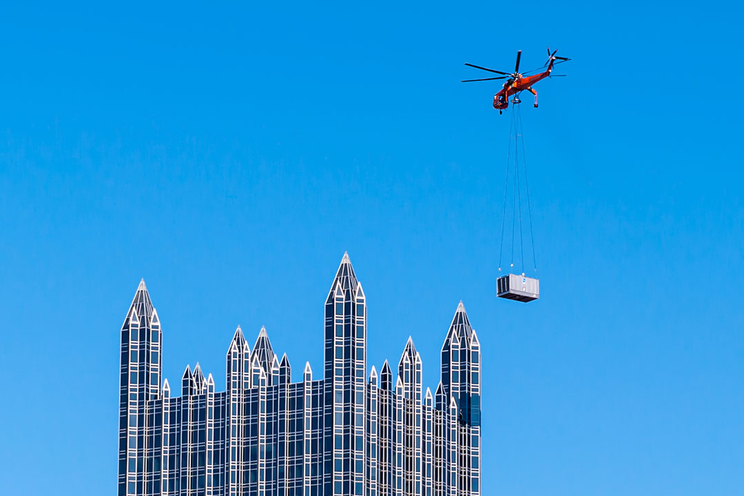 A skycrane lifts an HVAC unit on top of a large building.