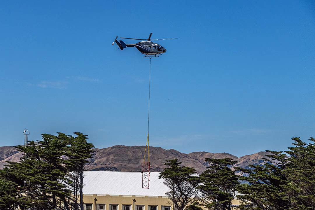 Helicopter rising after attaching radio mast component and lifting component clear of building and trees
