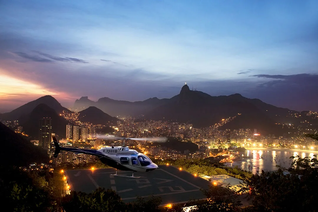 A helicopter departing a helipad overlooking Rio de Janeiro, illustrating terrain, obstacles, and visual complexity relevant to night flight operations.