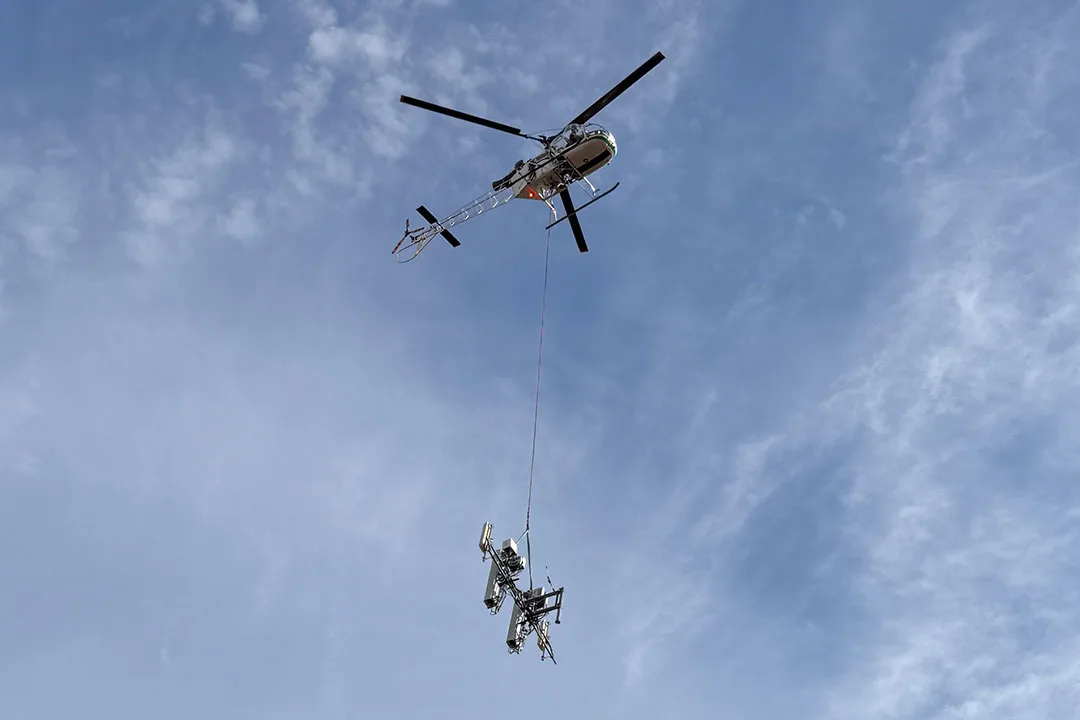 Helicopter performing an external load operation, lifting telecommunications equipment suspended on a long line against a clear blue sky.