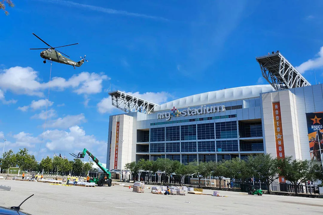 Heavy-lift helicopter rigging a suspended external load near a large stadium, demonstrating long-line precision placement in an urban infrastructure environment.