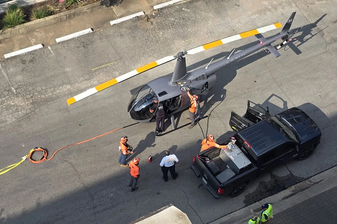 Overhead view of a helicopter on the ground during pre-lift staging, with crew members in high-visibility safety gear coordinating equipment and load preparation beside a support truck.