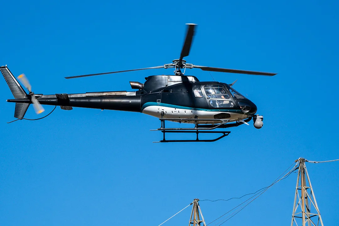 Light helicopter flying near distribution lines during close-range powerline inspection.