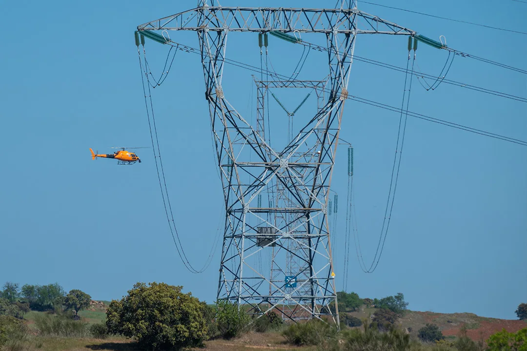 Helicopter inspecting high-voltage transmission tower in rural terrain.