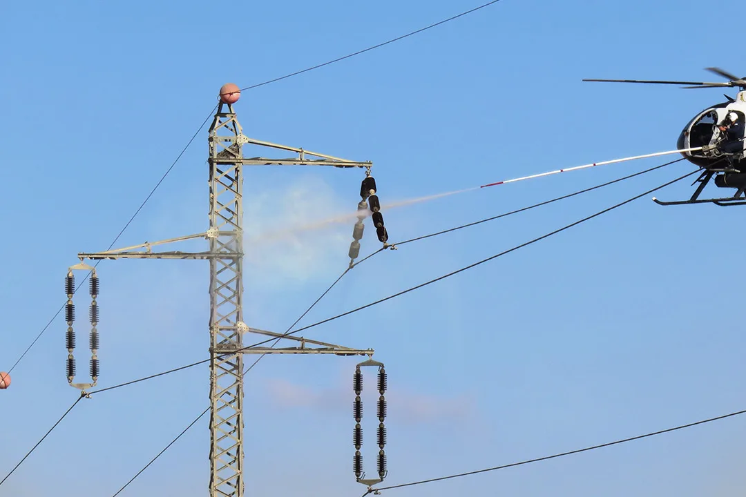 Helicopter performing live-line insulator washing on high-voltage transmission structure.