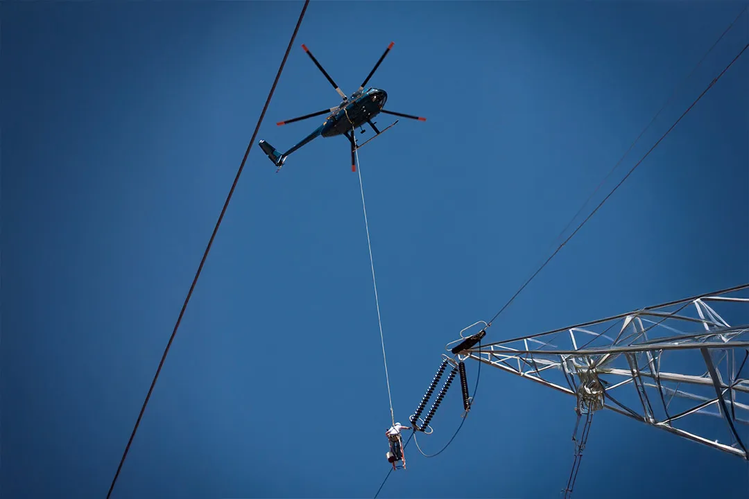Helicopter performing precision long-line work on energized transmission tower hardware.