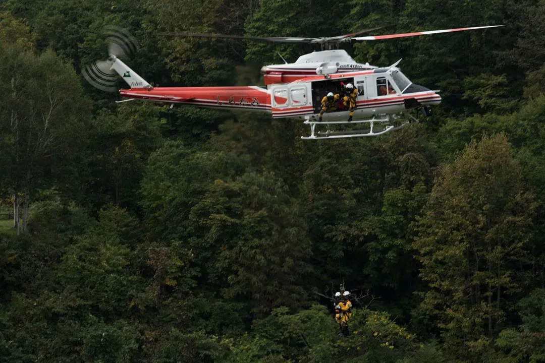 A Bell 412 helicopter hovers over a dense green forest while performing a precision winch rescue operation with two specialists suspended below the aircraft. | Photo: bellflight.com