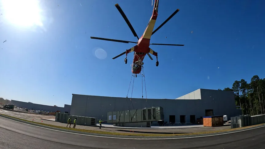 A Skycrane Helicopter begins lifting a massive rooftop HVAC unit off the ground to its destination on the roof.