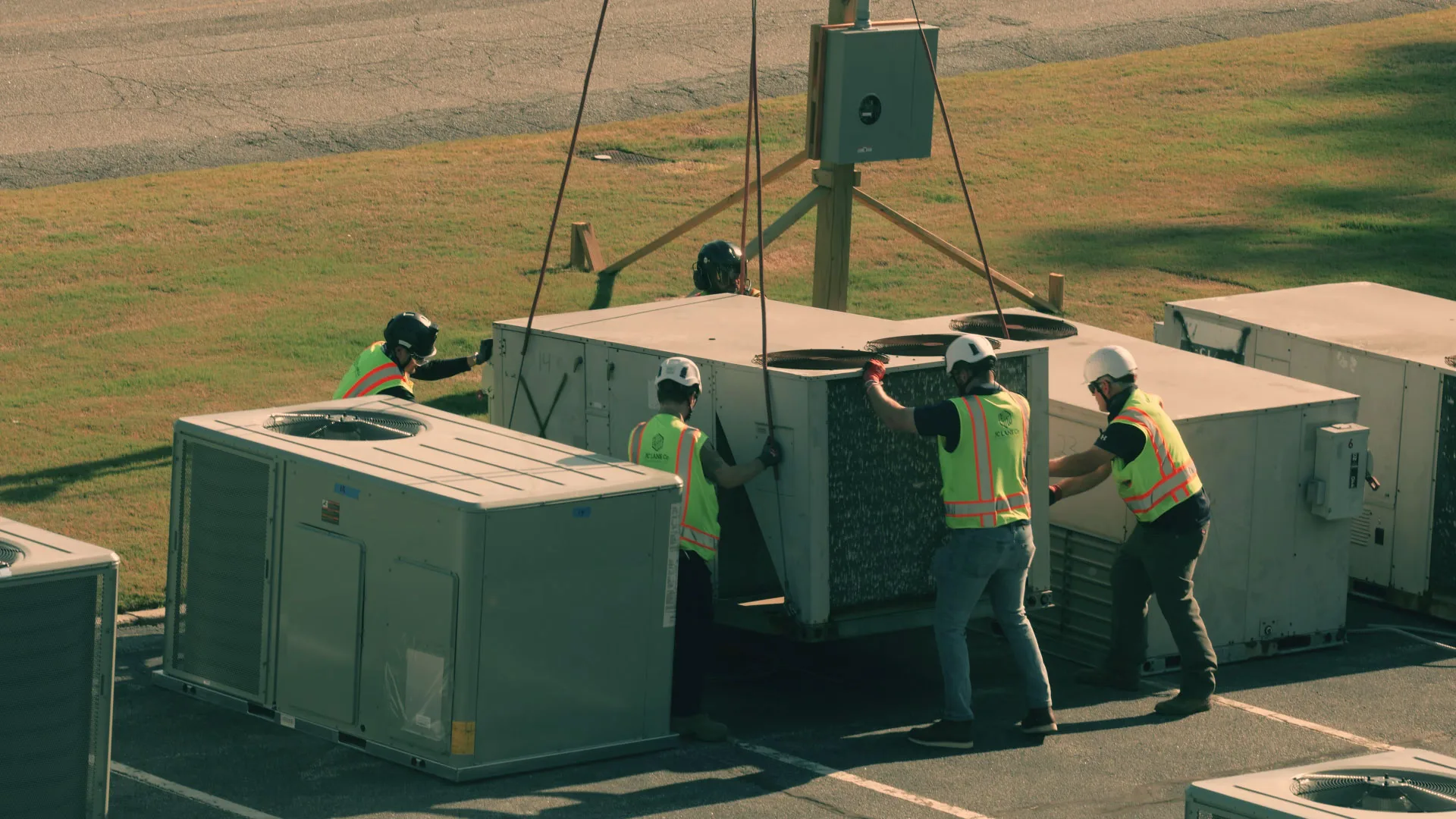 Four ground crew technicians guiding a heavy commercial air conditioning unit during a lift.