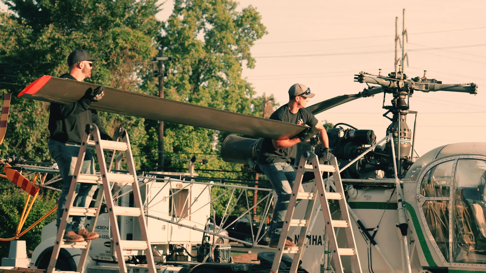 Two crew members lifting and carrying a helicopter rotor blade during assembly on a trailer.