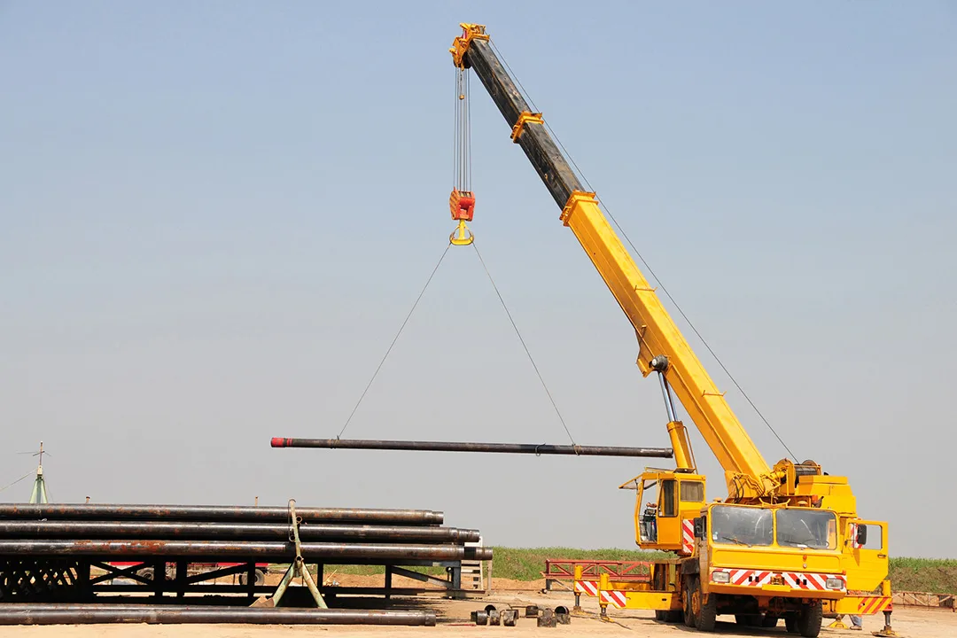 Yellow mobile crane lifting a long steel pipe using a two-point rigging configuration.