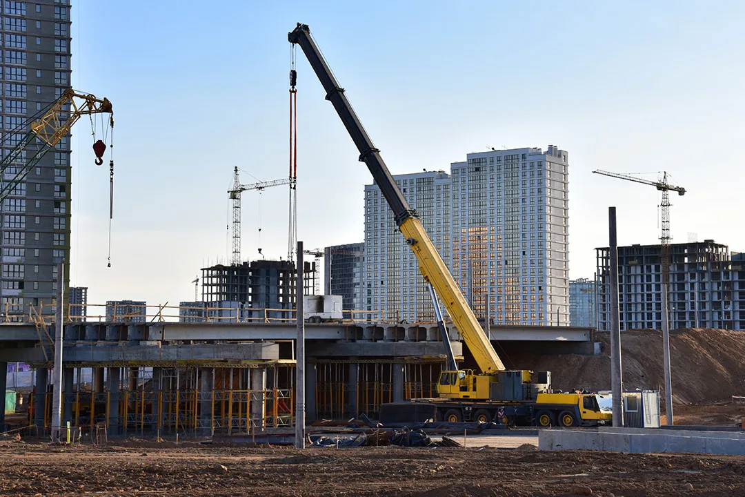 Mobile crane with an extended telescopic boom operating at an urban construction site surrounded by high-rise buildings.